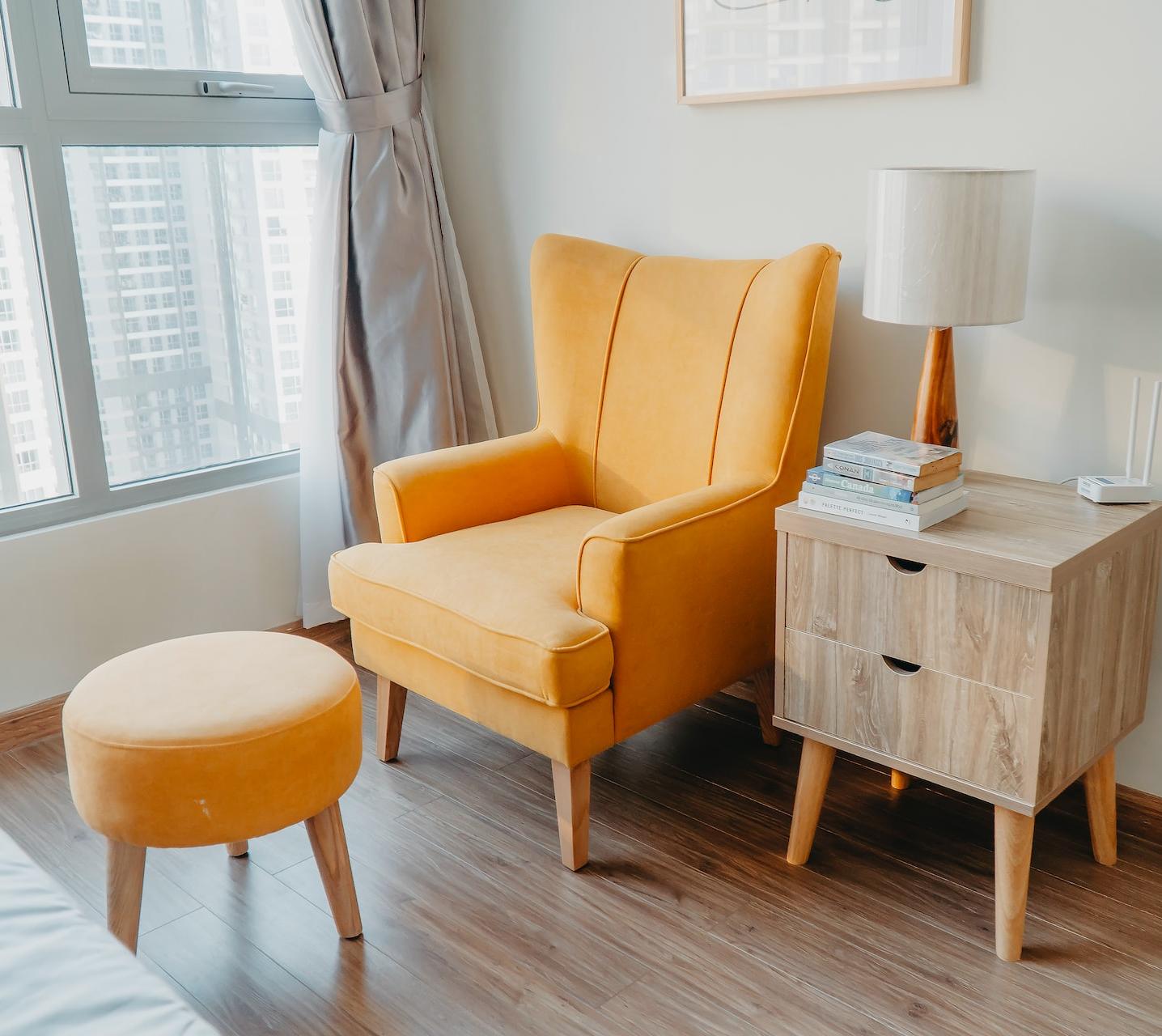 yellow armchair and stool beside wooden nightstand by the wall near glass window and bed